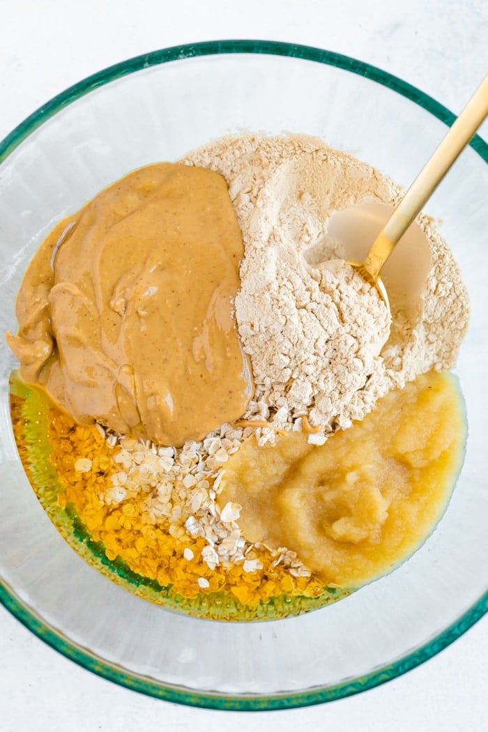 Glass mixing bowl with ingredients for peanut butter and jelly protein bites including apple sauce, oats, protein powder, peanut butter, and honey.