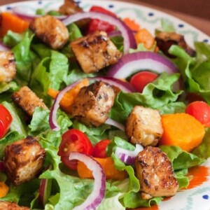 A bed of salad greens with red onion slices and maple lemon tempeh cubes on a dinner plate.