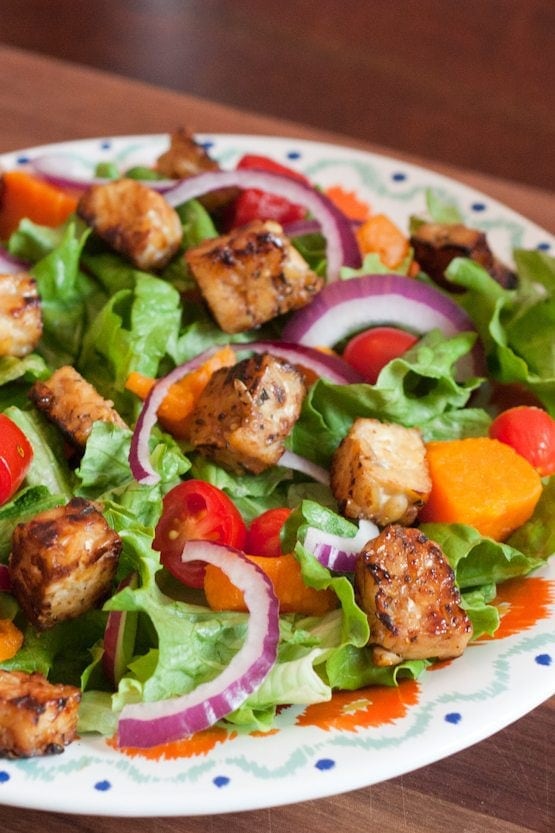 A bed of salad greens with red onion slices and maple lemon tempeh cubes on a dinner plate.