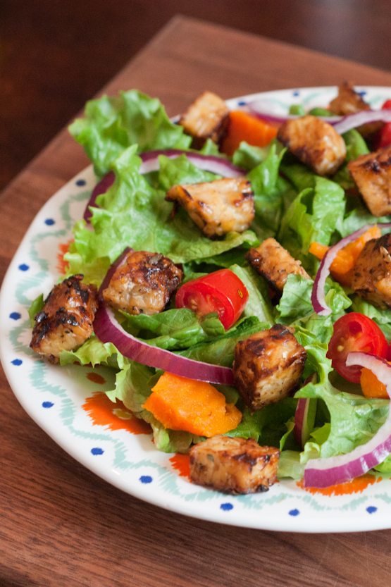 A bed of salad greens with red onion slices and maple lemon tempeh cubes on a dinner plate.