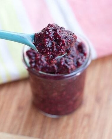 Mixed Berry Chia Seed Jam served in a small clear glass jar with light blue serving spoon on wood cutting board.