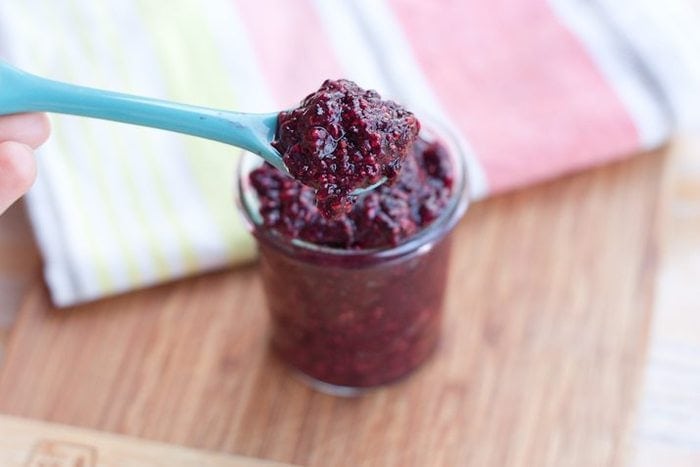 Mixed Berry Chia Seed Jam served in a small clear glass jar with light blue serving spoon on wood cutting board.