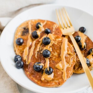 Three blueberry protein pancakes on a plate drizzled with peanut butter. Gold fork is also sitting on the plate.