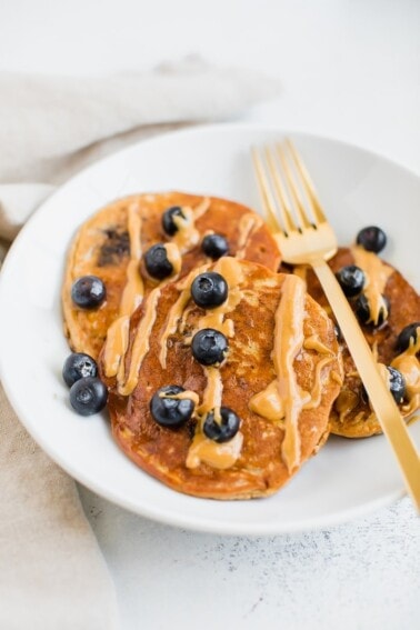 Three blueberry protein pancakes on a plate drizzled with peanut butter. Gold fork is also sitting on the plate.