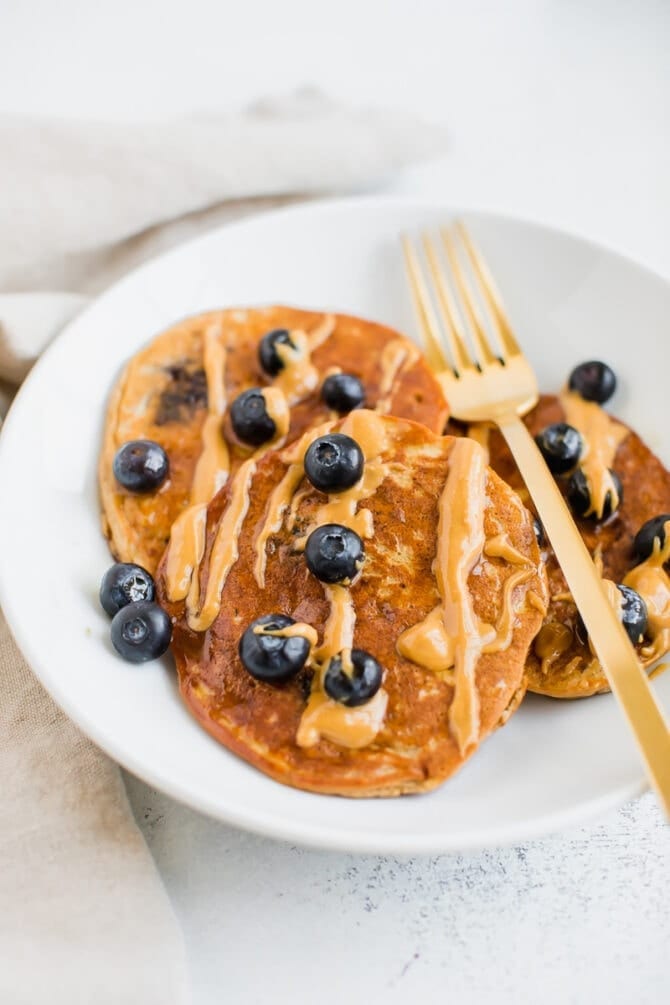 Three blueberry protein pancakes on a plate drizzled with peanut butter.