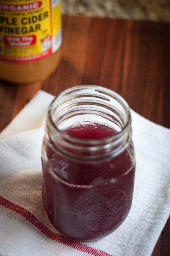 Mason jar with purple liquid sitting on a white towel. Jar of apple cider vinegar out of focus in the back of the image. 