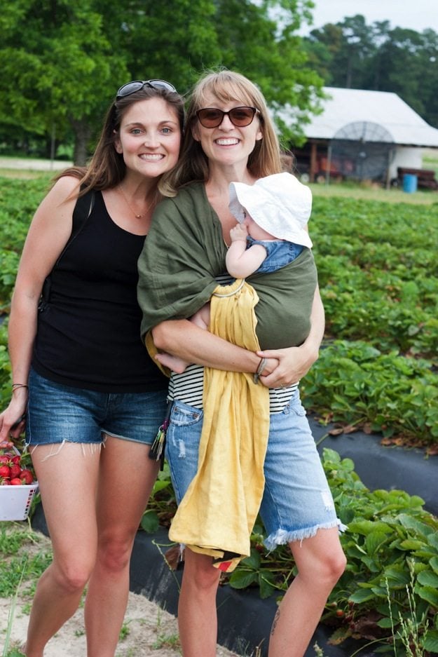 Two women looking at the camera smiling, one of the women is carrying a baby in a wrap across her body.