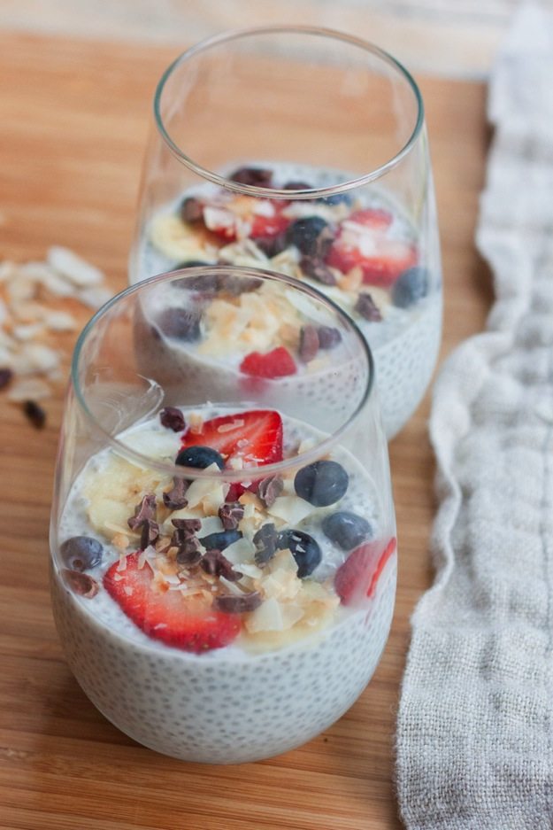 Two glasses with chia pudding, topped with coconut, banana, strawberries, blueberries and cacao nibs.