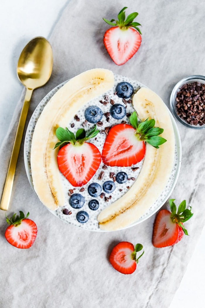 Bowl with chia pudding topped with a split banana, strawberries, blueberries and cacao nibs. A gold spoon, slices strawberries, and small bowl of cacao nibs are beside the bowl.