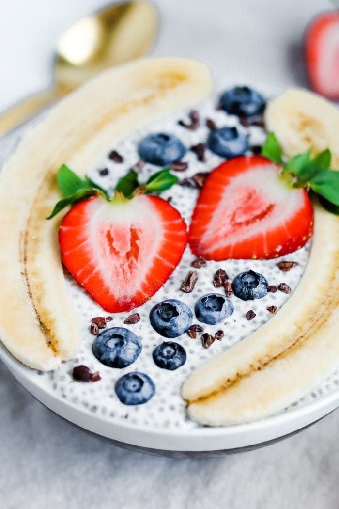 Bowl with chia pudding topped with a split banana, strawberries, blueberries and cacao nibs.