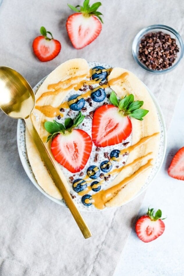 Bowl with chia pudding topped with a split banana, strawberries, blueberries and cacao nibs, and drizzled with peanut butter. A gold spoon, slices strawberries, and small bowl of cacao nibs are beside the bowl.