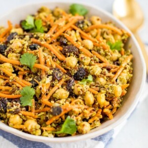 A carrot salad with raisins, quinoa, chickpeas in a bowl with a gold spoon on the right.