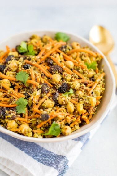 A carrot salad with raisins, quinoa, chickpeas in a bowl with a gold spoon on the right.