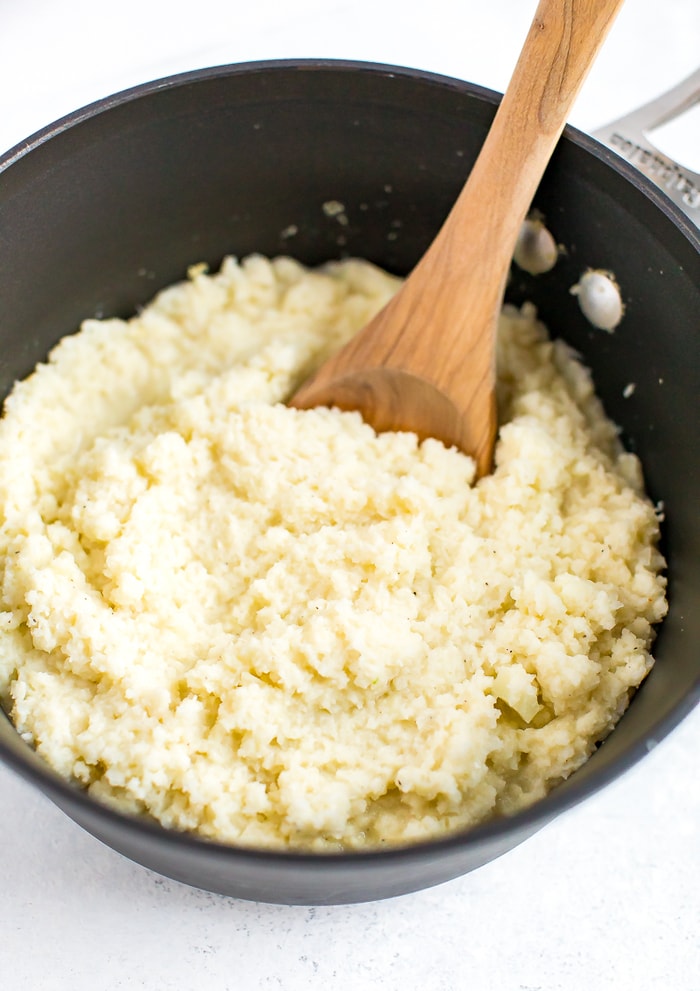 Pot with a wooden spoon stirring cauliflower cheese grits.