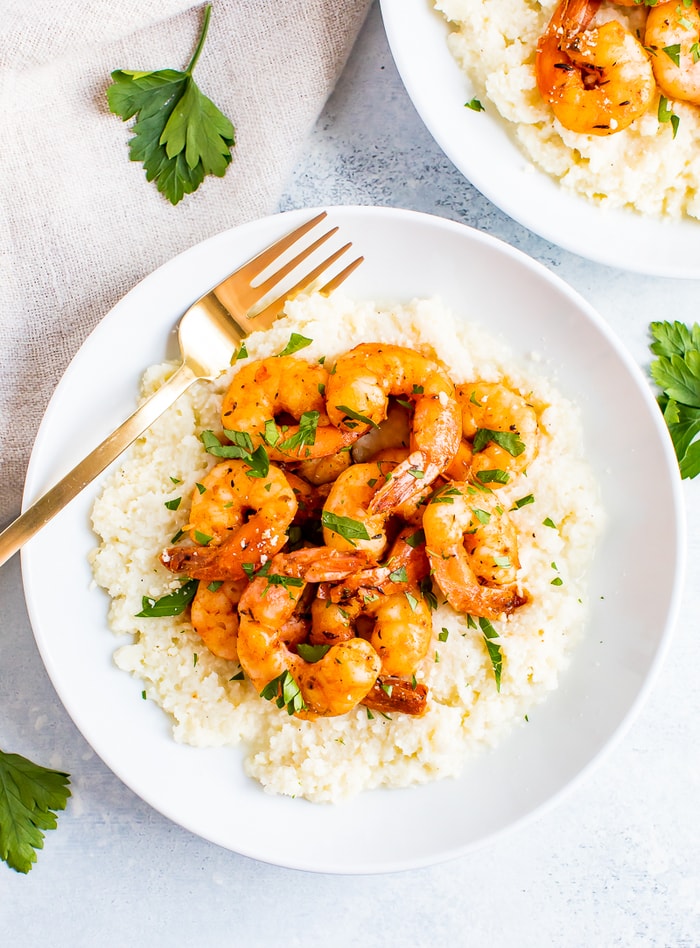 Plate of cauliflower cheese grits topped with blackened shrimp and chipped fresh parsley. A fork is resting on the plate.