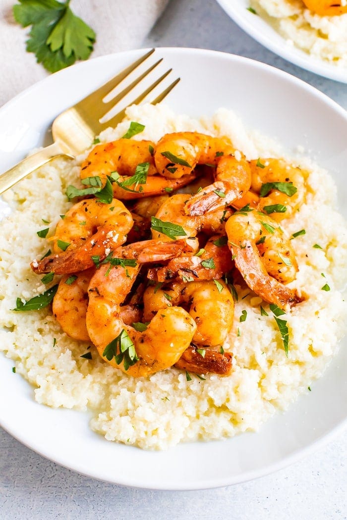 Plate of cauliflower cheese grits topped with blackened shrimp and chipped fresh parsley. A fork is resting on the plate.