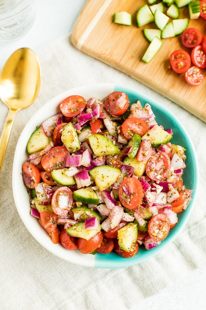 Cucumber tomato salad in a bowl with gold spoon on the left and cutting board on the right. 