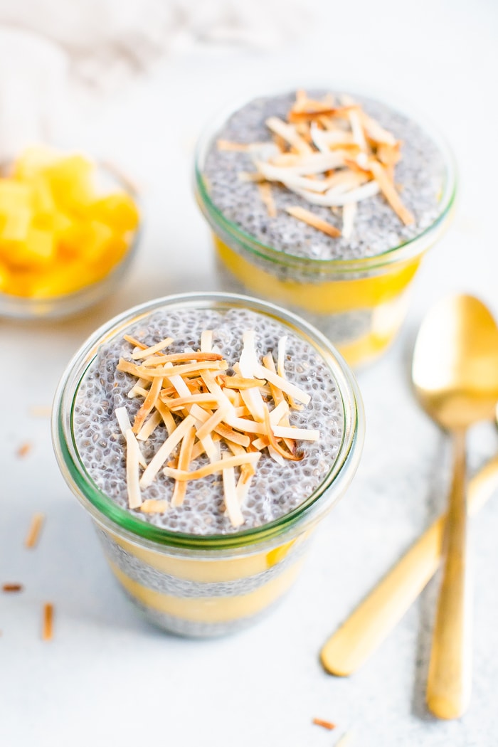 Two jars of coconut mango chia pudding topped with toasted coconut flakes. Jars are surrounded by two spoons, a napkin and mango chunks.