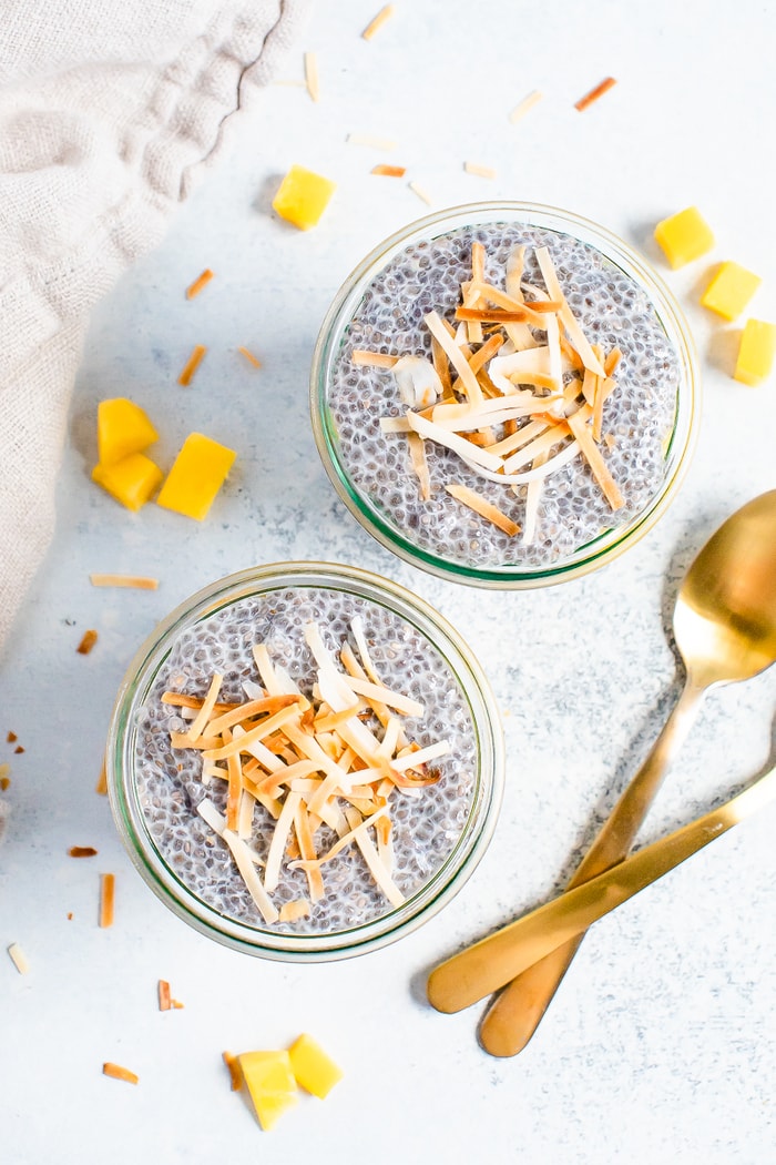 Two jars of coconut mango chia pudding topped with toasted coconut flakes. Jars are surrounded by two spoons, a napkin and mango chunks.