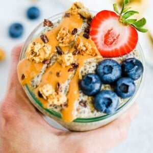 Hand holding a jar of peanut butter chia pudding topped with peanut butter, cacao nibs, granola, a strawberry and blueberries.