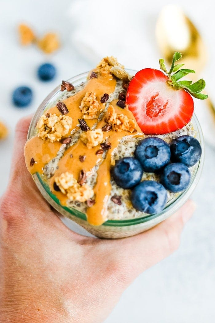 Hand holding a jar of peanut butter chia pudding topped with peanut butter, cacao nibs, granola, a strawberry and blueberries.