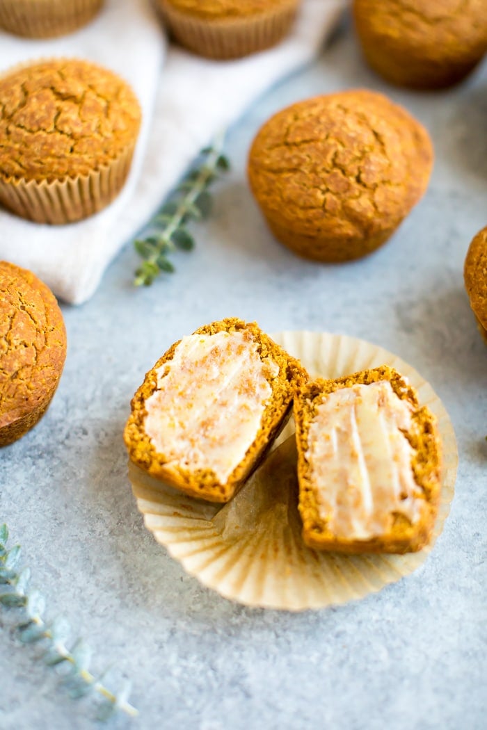 Healthy pumpkin cornbread muffins on a table with a cloth napkin and greenery sprigs. One muffin is sliced and spread with ghee.