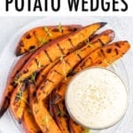 Plate of grilled sweet potato wedges on a plate, topped with fresh herbs. A creamy dipping sauce is to the side.