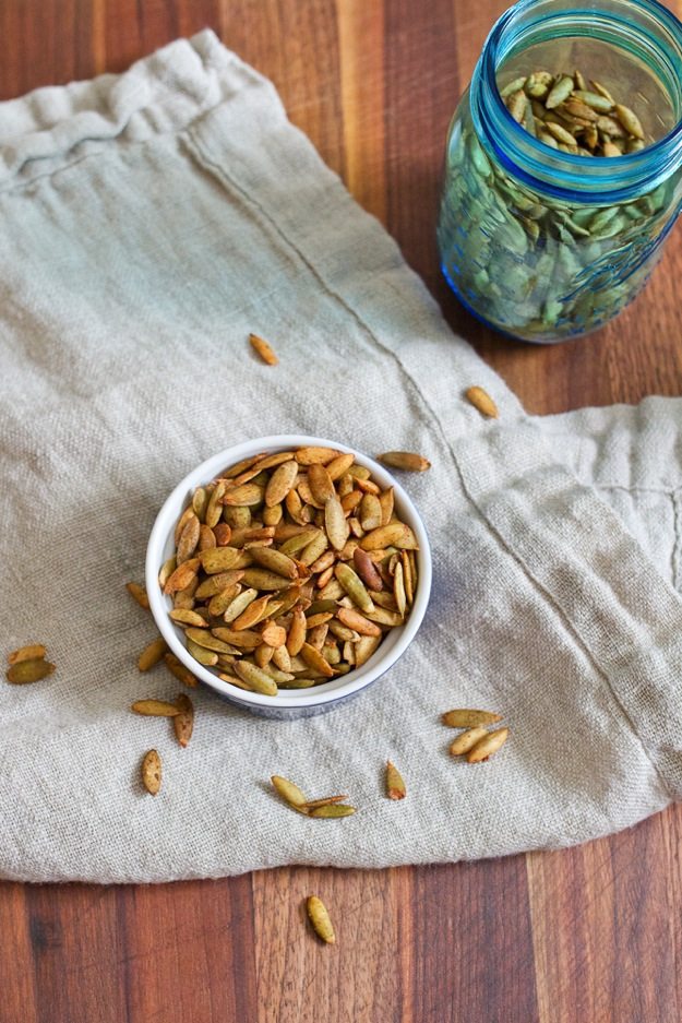 Pumpkin Spiced Roasted Pepitas in a small dish. The dish is resting on a linen dish cloth and there is a blue jar of additional pepitas nearby.