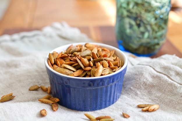 Pumpkin Spiced Roasted Pepitas in a shallow bowl placed on a linen dish cloth. There are additional pepitas scattered around the dish on the towel and a blue jar of extra pepitas in the background.