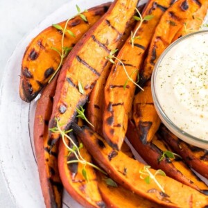 Plate of grilled sweet potato wedges on a plate, topped with fresh herbs. A creamy dipping sauce is to the side.