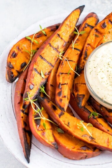 Plate of grilled sweet potato wedges on a plate, topped with fresh herbs. A creamy dipping sauce is to the side.