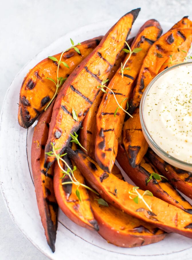 Plate of grilled sweet potato wedges on a plate, topped with fresh herbs. A creamy dipping sauce is to the side.