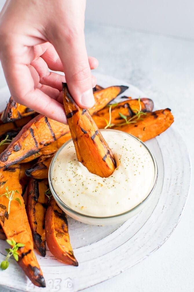 Hand dipping a grilled sweet potato wedge into a bowl of creamy dip.