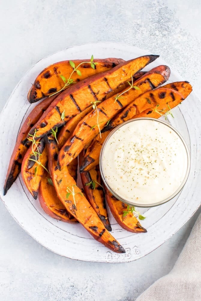 Plate of grilled sweet potato wedges on a plate, topped with fresh herbs. A creamy dipping sauce is to the side.