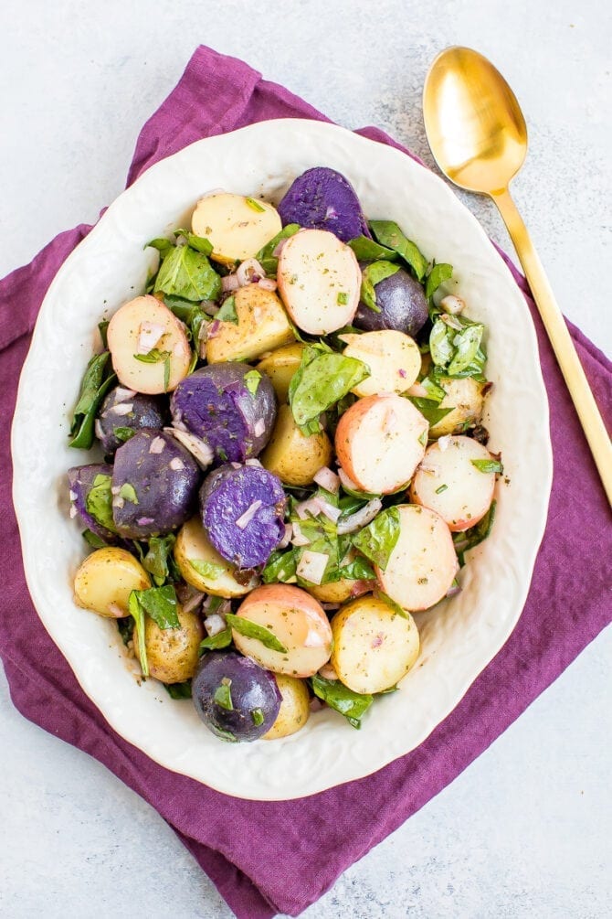 Tri-colored potato salad with shallots and spinach in a white bowl with a purple napkin and gold spoon. 