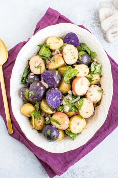 Tri-colored potato salad with shallots and spinach in a white bowl with a purple napkin and gold spoon.