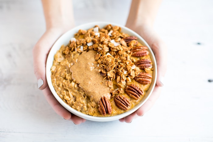 Hand holding a bowl of egg white protein pumpkin oatmeal topped with granola, peanut butter and pecans.
