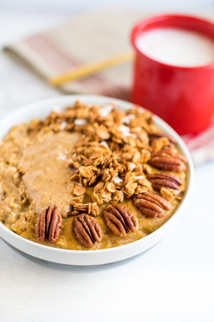 Protein Pumpkin Oatmeal with Egg Whites in a bowl, topped with granola, almond butter and pecans. A mug of almond milk to the side.