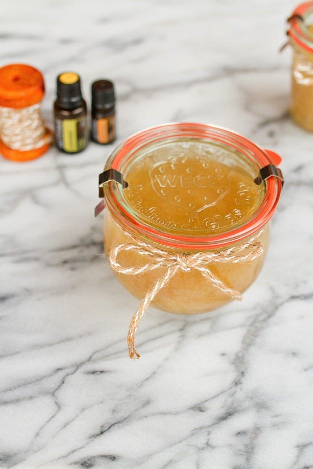 Small glass jar filled with sugar scrub, twine bow wrapped around top of jar sitting on a marble countertop. Essential oil bottles in background.