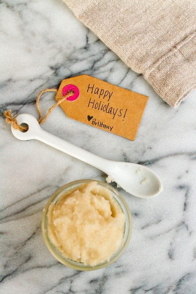 Small jar of sugar scrub, white scooping spoon and gift tag that reads 'Happy Holidays!' sitting on marble countertop.