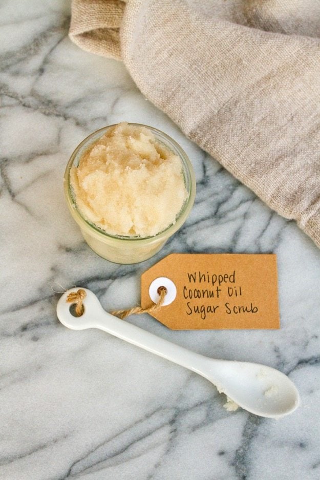 Small mason jar filled with sugar scrub sitting on marble counter with linen towel and scoop with a gift tag that reads 'Whipped Coconut Oil Sugar Scrub'