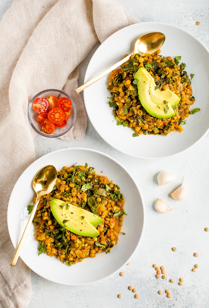 Two bowls of lentil stew with kale and topped with avocado slices. Gold spoons are on the sides of the bowls, and a bowl of cheery tomatoes are around the bowls along with garlic and a cloth napkin.