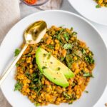 Bowl with lentil stew topped with avocado slices. A gold spoon is on the bowl. A bowl of sliced cherry tomatoes is to the side.