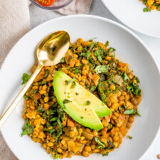Bowl with lentil stew topped with avocado slices. A gold spoon is on the bowl. A bowl of sliced cherry tomatoes is to the side.