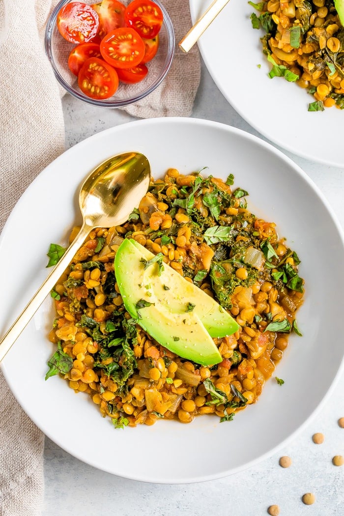 Bowl with lentil stew topped with avocado slices. A gold spoon is on the bowl. A bowl of slices cherry tomatoes is to the side.