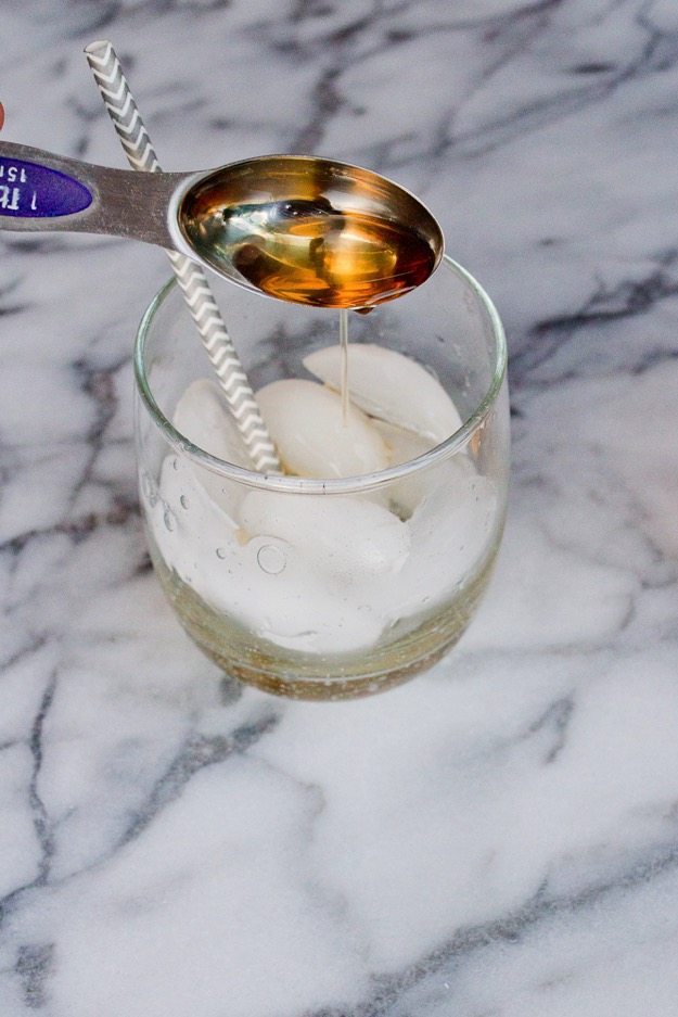 Teaspoon of vinegar poured into a Sparkling Elderflower Apple Lime Vinegar Mocktail in a clear glass on white marble countertop.