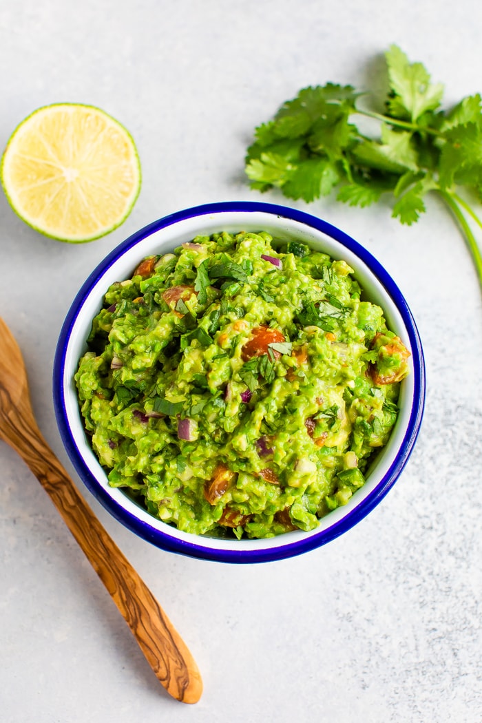 Bowl of guacamole on a table with a wood spoon, lime and cilantro.