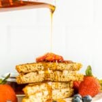 Pyrex measuring cup pouring maple syrup over two slices of stuffed french toast on a plate with berries.