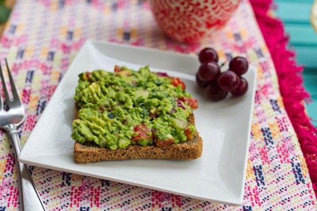 Guacamole spread over a slice of toast on a white plate. A cluster of grapes is also on the plate.