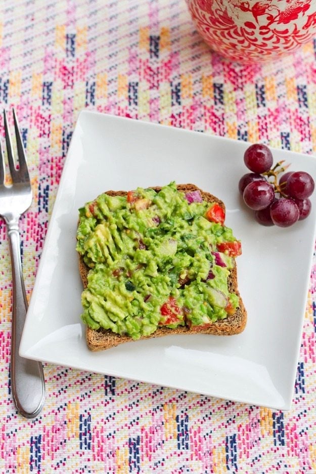 Guacamole spread over a slice of toast on a white plate. A cluster of grapes is also on the plate.
